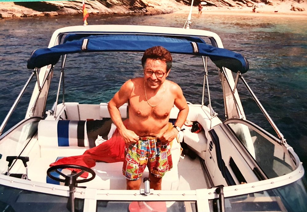 José Luis Gimeno sonriente en bañador de colores en la cubierta de su lancha, fondeada en una cala
